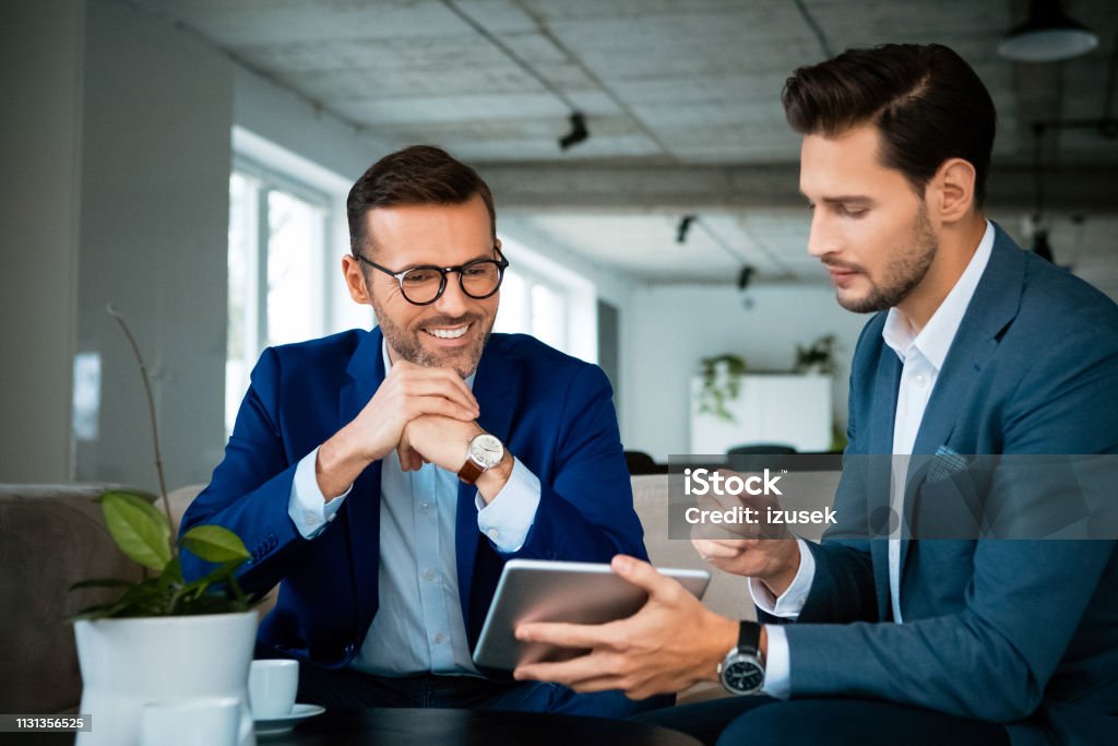 Smiling colleagues discussing over digital tablet Smiling colleagues planning strategy during meeting. Confident male professionals are discussing over digital tablet. They are working at creative office. Discussion Stock Photo