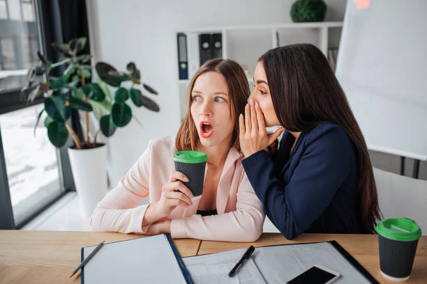 dos jóvenes empresarias se sientan juntas en la habitación de la mesa. morena susurra a oreja otra hembra. chismean. chica rubia mantener una taza de café. - cotilleo fotografías e imágenes de stock