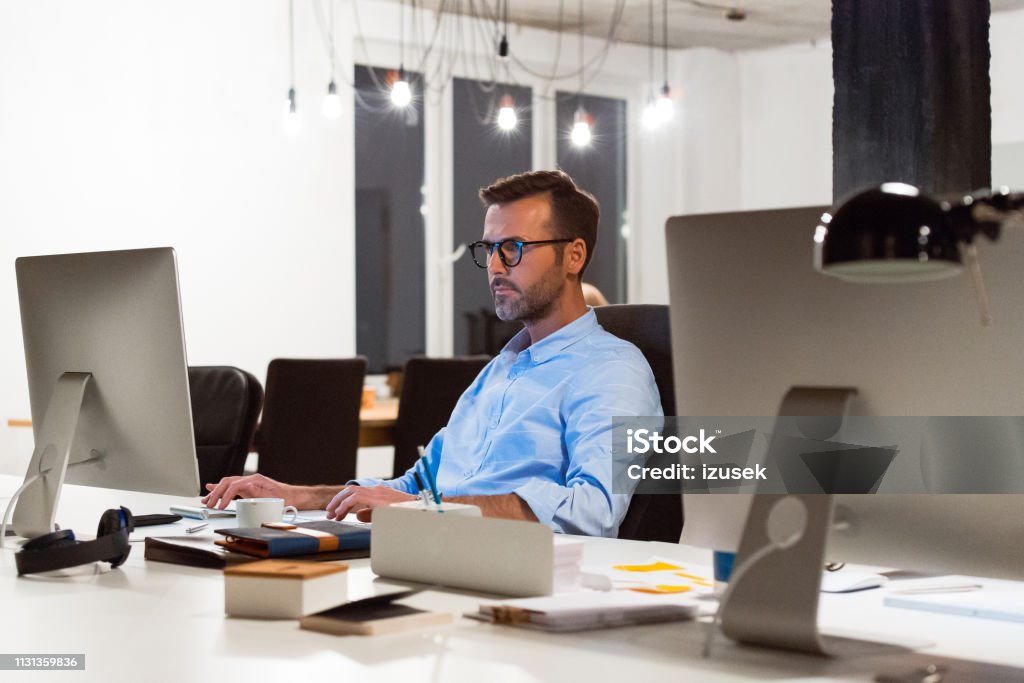 Confident businessman using computer at desk Confident mature entrepreneur using computer at creative workplace. Businessman is working late at office. Design professional is sitting at desk. Creative Occupation Stock Photo