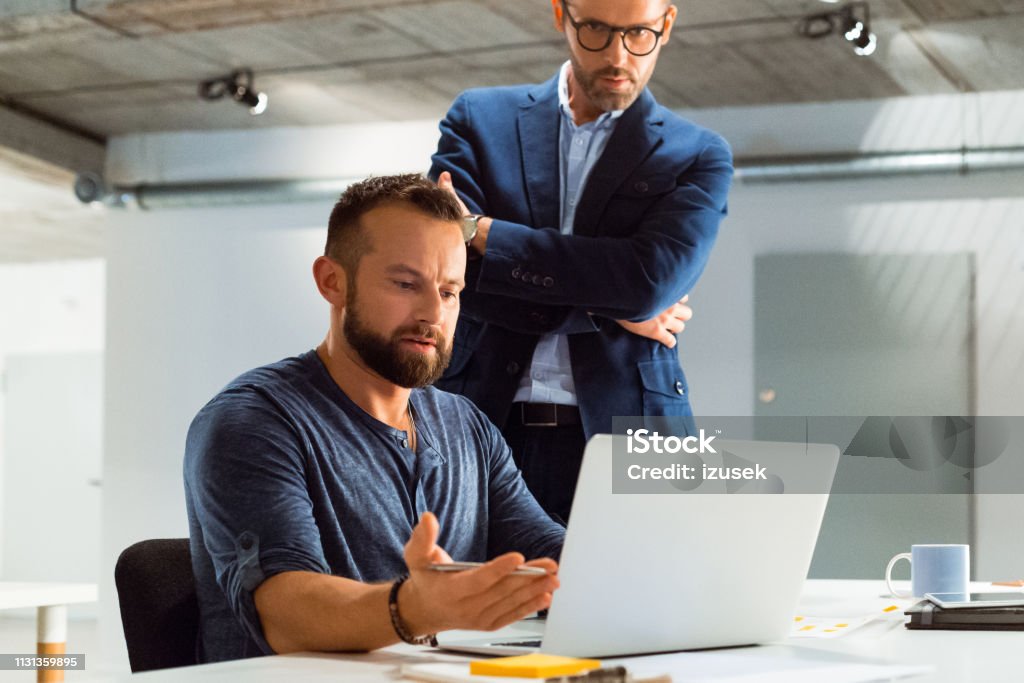 Businessman discussing over laptop with colleague Confident businessman discussing over laptop with colleague during meeting. Male entrepreneurs are working late at office. They are planning strategy at creative workplace. Computer Stock Photo