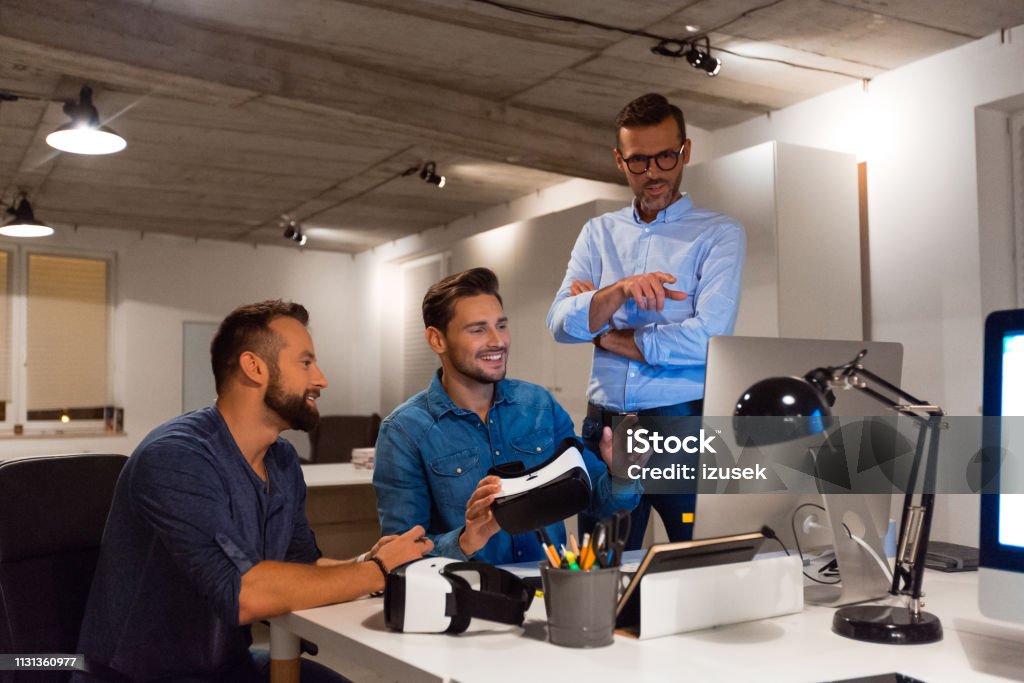 Male professionals discussing over computer Smiling businessman holding virtual reality headset sitting amidst colleagues. Male professionals are discussing over computer at creative office. Entrepreneurs are working late. Office Stock Photo