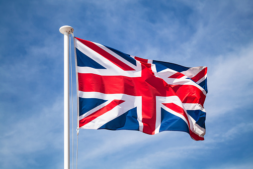The national flag of the United Kingdom is the Union Jack, also known as the Union Flag waving on wind over blue sky background.
