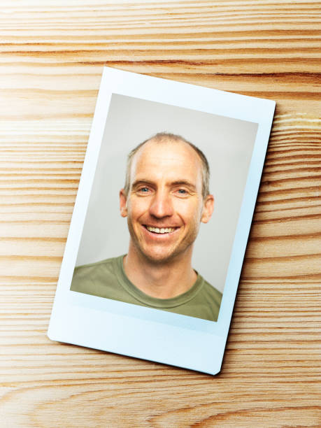 Printed portrait from instant camera of mid adult caucasian man with happy expression looking at camera against gray white background. Vertical shot of american real people smiling in studio with short balding hair and blue eyes. (Fuji Instax film). Photography from a DSLR camera. Sharp focus on printed photo.