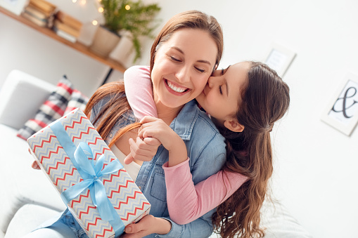 Young woman and girl at home celebrating mother's day sitting on sofa daughter hugging mom holding present box kissing cheek closed eyes pleasure close-up.