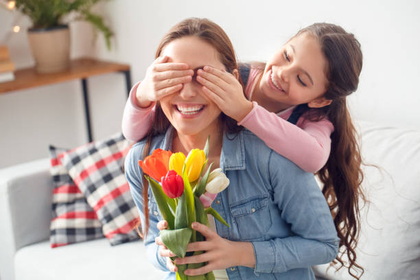 Mother and daughter at home mother's day daughter covering eyes of mother sitting with flowers Young woman and girl at home celebrating mother's day daughter covering eyes of mother sitting holding bouquet of flowers laughing cheerful close-up cute pregnant woman with gift stock pictures, royalty-free photos & images