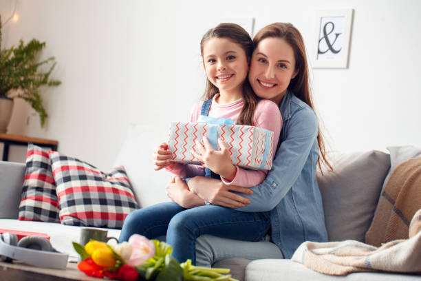 Mother and daughter at home birthday sitting mother hugging daughter holding gift box smiling happy Young woman and girl at home celebrating birthday daughter sitting on mom's knees holding gift box mother hugging her smiling happy cute pregnant woman with gift stock pictures, royalty-free photos & images