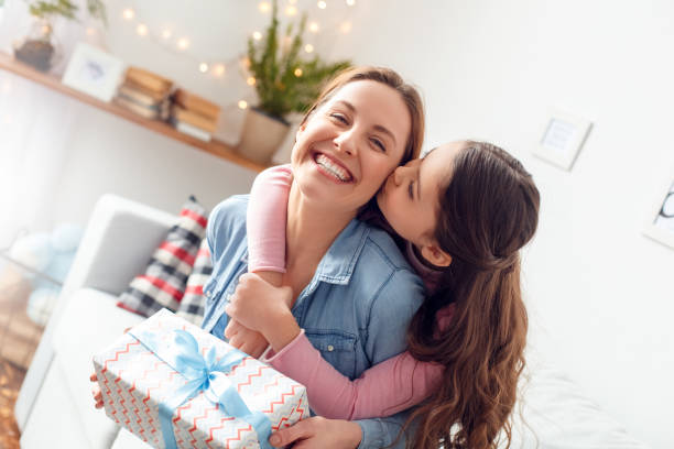 Mother and daughter at home mother's day sitting daughter hugging mom kissing cheek joyful Young woman and girl at home celebrating mother's day sitting on sofa daughter hugging mother kissing cheek mom laughing joyful holding gift box kissing photos stock pictures, royalty-free photos & images