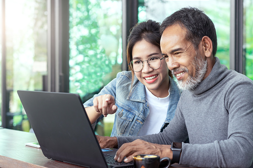 Attractive mature asian man with white stylish short beard looking at laptop computer with teenage eye glasses hipster woman in cafe. Teaching internet online or wifi technology in older man concept.