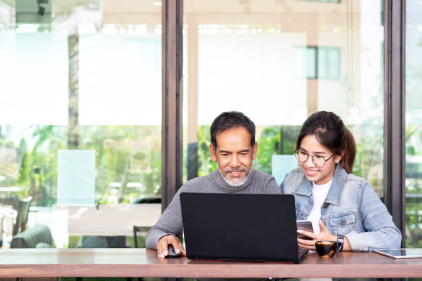 homem asiático maduro atrativo com a barba curta à moda branca que olha o computador portátil com óculos de olho adolescente mulher do hipster no café. ensinando a tecnologia em linha do internet ou do wifi no conceito mais idoso do homem. - togetherness customer bonding business - fotografias e filmes do acervo