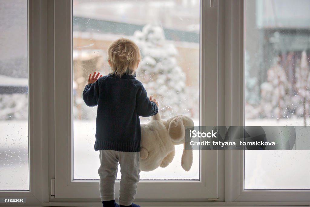 Kleinkind steht vor einer großen französischen Tür, lehnt sich dagegen und schaut nach draußen auf eine schneebedeckte Natur - Lizenzfrei Winter Stock-Foto