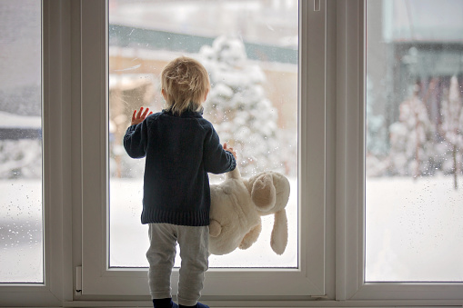 Toddler child standing in front of a big french doors, leaning against it looking outside at a snowy nature, holding teddy toy