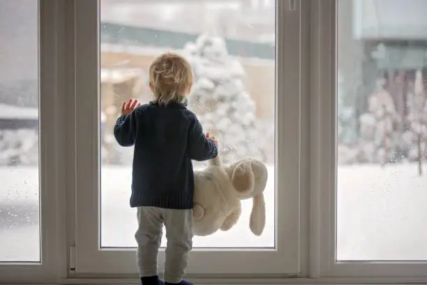 Photo of Toddler child standing in front of a big french doors, leaning against it looking outside at a snowy nature