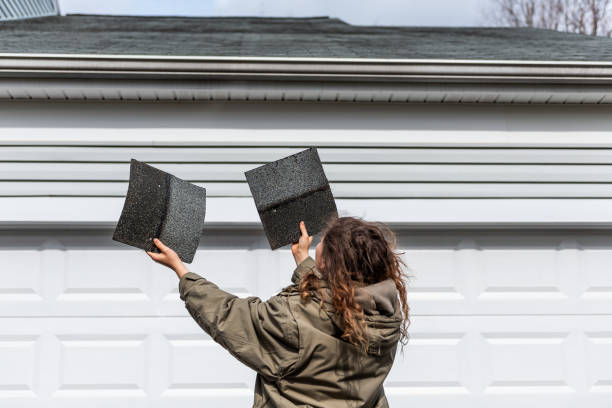 Young woman female homeowner standing in front of house garage in coat jacket during winter storm holding two roof tile shingles inspecting damage Young woman female homeowner standing in front of house garage in coat jacket during winter storm holding two roof tile shingles inspecting damage replacement stock pictures, royalty-free photos & images