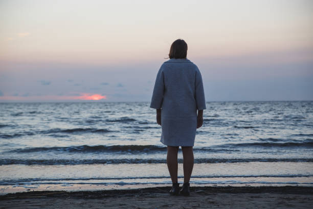 istock Woman standing alone on the beach under cloudy sky from behind 1131406098