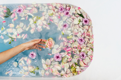 Top view of bath filled with blue bubble water, flowers and petals with woman's hand, spa or selfcare concept