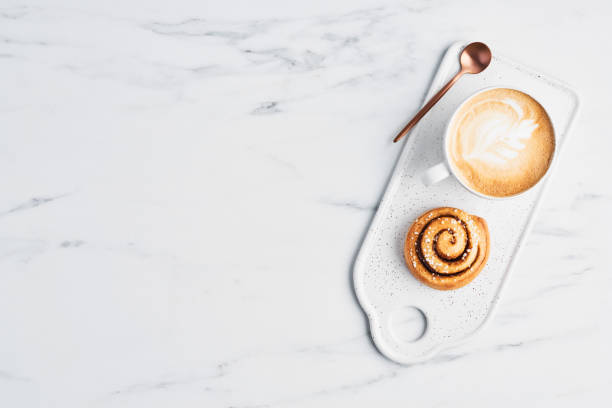 Freshly baked cinnamon roll and coffee with latte art Freshly baked cinnamon roll with spices and cocoa filling and coffee or cappuccino with latte art on white serving plate over white marble background. Top view. Copy space for text. Swedish breakfast. cafeteria photos stock pictures, royalty-free photos & images
