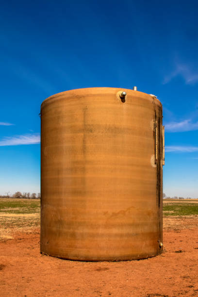 primo piano del vecchio serbatoio d'acqua arrugginito seduto in pascolo con terra rossa calpestata intorno ad esso su un terreno pianeggiante con linea di alberi in lontananza e bellissimo cielo blu nell'oklahoma occidentale - rusty storage tank nobody photography foto e immagini stock