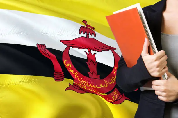 Photo of Learning Bruneian language concept. Young woman standing with the Brunei flag in the background. Teacher holding books, orange blank book cover.