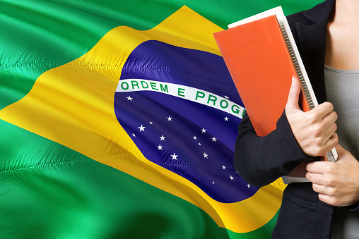 Learning Brazilian language concept. Young woman standing with the Brazil flag in the background. Teacher holding books, orange blank book cover.
