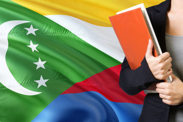 Learning Comoran language concept. Young woman standing with the Comoros flag in the background. Teacher holding books, orange blank book cover.