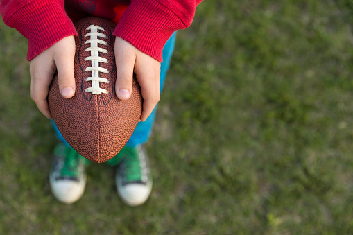 Top view on hands of little kid boy holding football on the stadium on a sunny day. Child  ready to throw a football. Sport concept. Sport activities for children outdoors.
