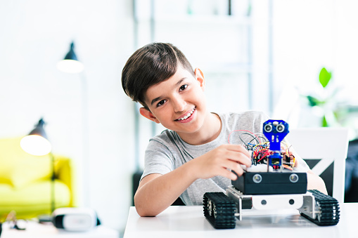 Cheerful smart schoolboy sitting at the table and constructing a robotic device at home