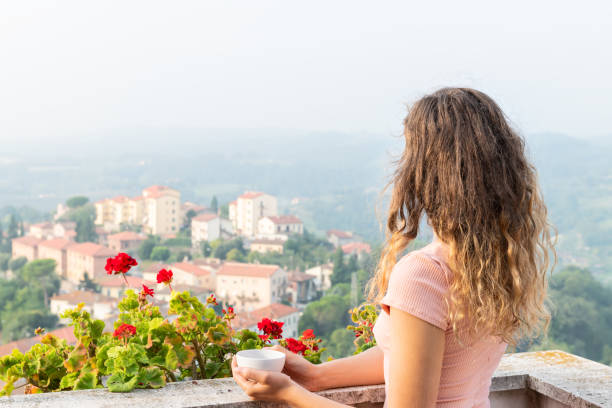 donna sulla terrazza con fiori di geranio rosso fuori in italia con vista sulle montagne di chiusi paesaggio urbano umbria vicino alla toscana che beve tè o caffè dalla tazza a colazione mattina con nebbia nebbia - balcony women patio coffee foto e immagini stock