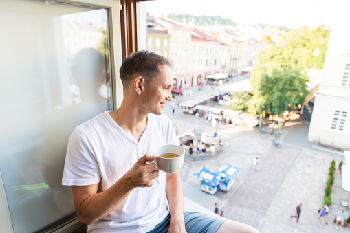 Young happy man sitting on windowsill holding tea cup in apartment room with view of Lviv, Ukraine old market town square in morning