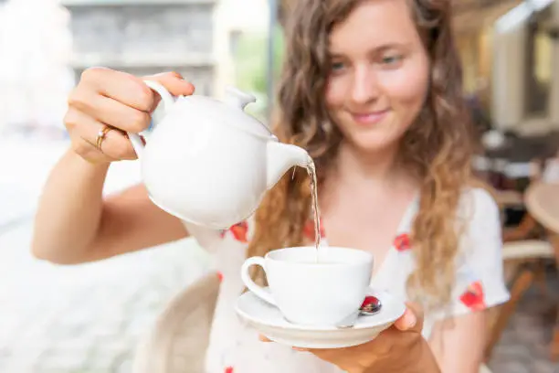 Young girl woman sitting in European outdoor cafe restaurant pouring green tea in white cup in summer in Lviv or Lvov, Ukraine city