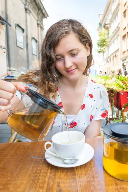 Young girl woman sitting in European outdoor cafe restaurant pouring green tea in cup in summer in Lviv or Lvov, Ukraine city