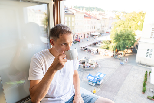 Young man sitting on windowsill with teacup holding drinking tea cup in apartment room with view of Lviv, Ukraine old market town square in morning