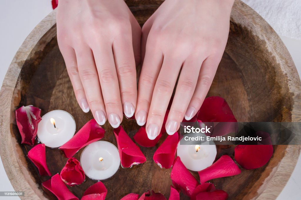 Female hands with french nails polish style and wooden bowl with water and floating candles and red rose petals Beautiful female hands with french nails polish style and wooden bowl with water and floating candles and red rose petals. Manicure and Beauty concept. Close up, selective focus Adult Stock Photo