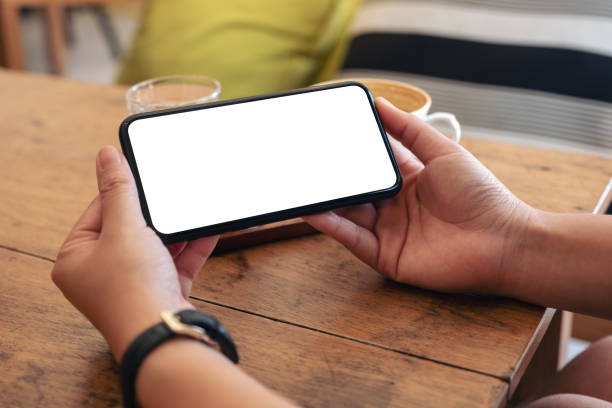 istock Mockup image of hands holding black mobile phone with blank screen horizontally with coffee cup on wooden table in cafe 1131526778