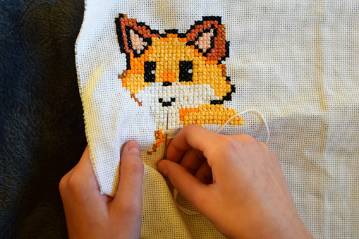 Cross stitching embroidery of cute little fox in progress in human hands. Teen girl hands sewing with needle and colorful threads. Handicraft hobby concept.