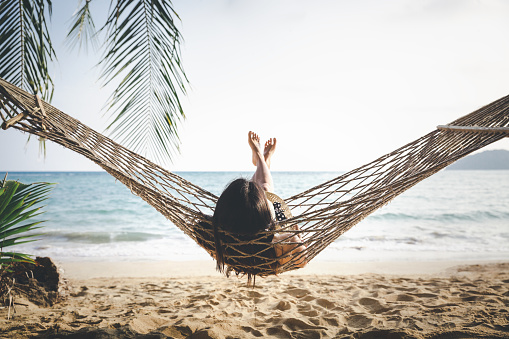 Summer vacations concept, Happy woman with white bikini, hat and shorts Jeans relaxing in hammock on tropical beach at sunset, Koh mak, Thailand