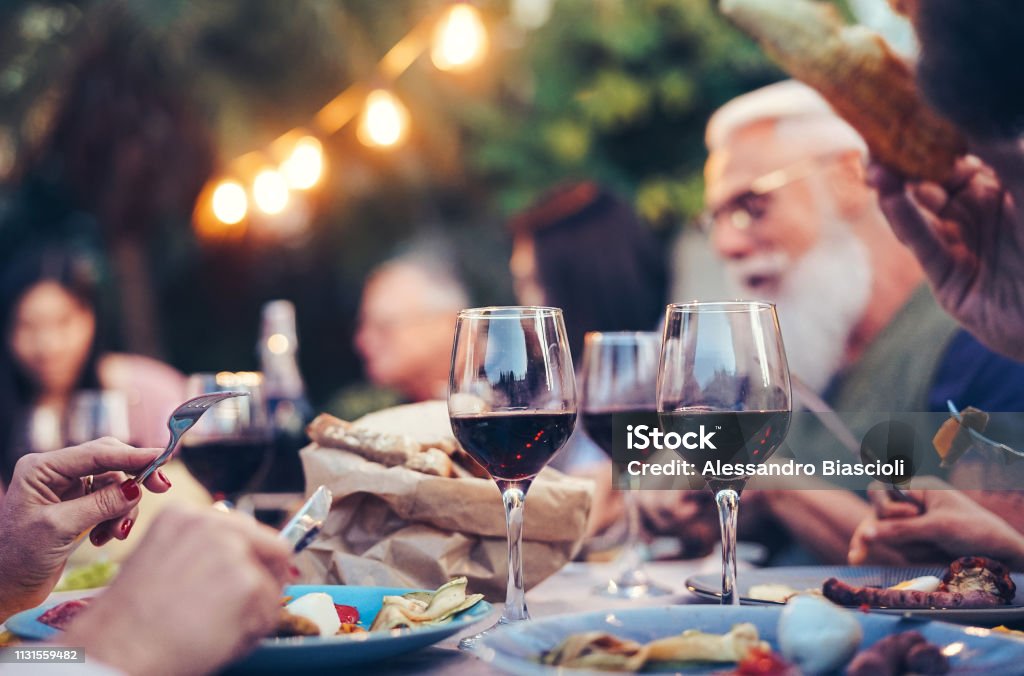 Feliz familia comiendo y bebiendo vino tinto en la cena barbacoa fiesta al aire libre-maduro y los jóvenes que cenan juntos en la azotea-actividades de fin de semana de los jóvenes y ancianos-enfoque en Wineglass - Foto de stock de Vino libre de derechos