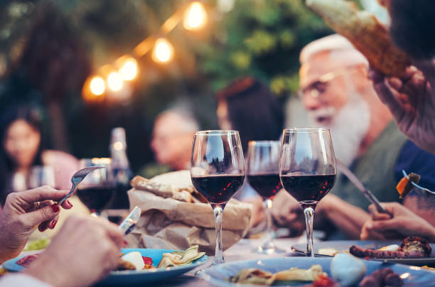 happy family eating and drinking red wine at dinner barbecue party outdoor - mature and young people dining together on rooftop - youth and elderly weekend lifestyle activities - focus on wineglass - dining senior adult friendship mature adult imagens e fotografias de stock
