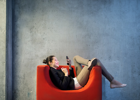 A young businesswoman with smartphone sitting on red armchair in office, a gray concrete wall in the background.