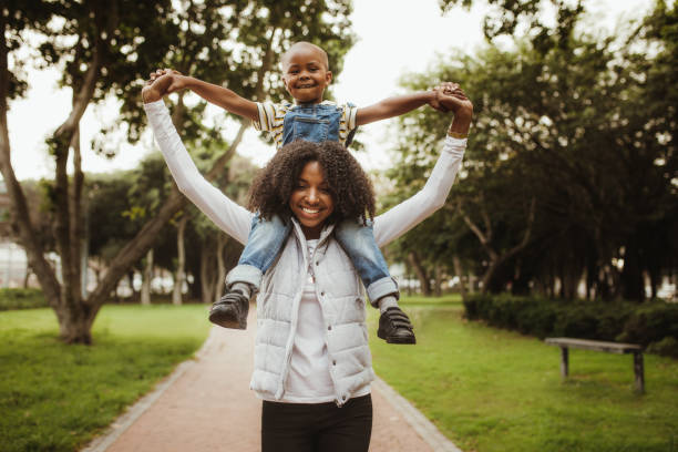 Happy mother giving son a piggyback ride on her shoulders in park. African woman carrying his boy on her shoulders while walking in a park.