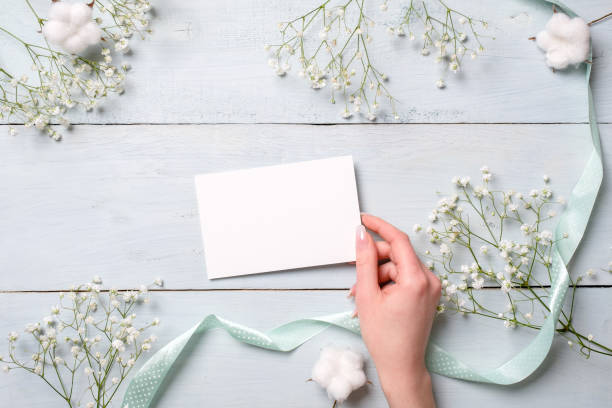 istock Womens hand holding blank paper card on light blue wooden background. Rustic table with flowers, wedding invitation card, letter, greeting card for Womens day. Trendy minimal design, Flat composition 1131568650