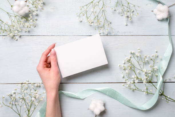 istock Womens hand holding blank paper card on light blue wooden background. Rustic table with flowers, wedding invitation card, letter, greeting card for Womens day. Trendy minimal design, Flat composition 1131568651