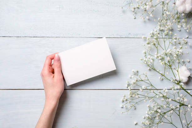 istock Womens hand holding blank paper card on light blue wooden background. Rustic table with flowers, wedding invitation card, letter, greeting card for Womens day. Trendy minimal design, Flat composition 1131568669