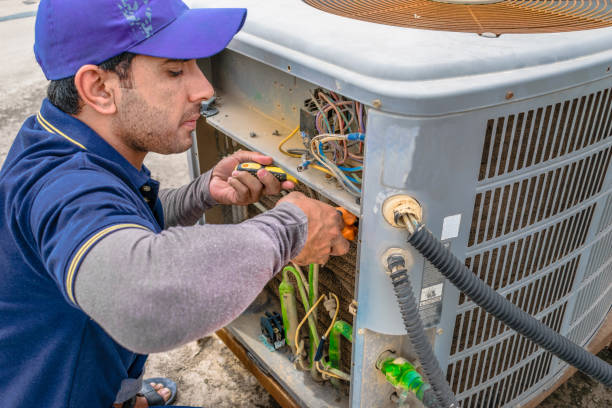 ein professioneller elektriker befestigt die schwere einheit einer klimaanlage an der dachterrasse eines gebäudes und trägt blaue uniform und kopfkappe - techniker stock-fotos und bilder