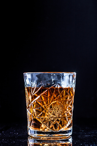 Drinking glass with whiskey on black background. Golden bourbon alcohol drink on table with copy space