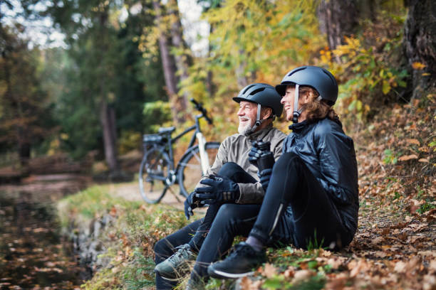 una pareja de ancianos con electrobicis sentado al aire libre en el parque en otoño, descansando. - cycling senior adult sports helmet men fotografías e imágenes de stock
