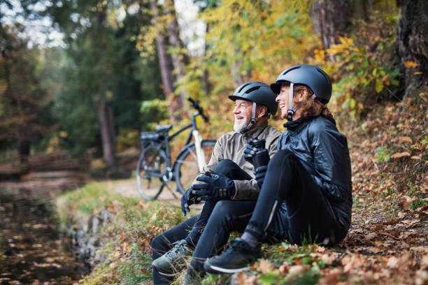 a senior couple with electrobikes sitting outdoors in park in autumn, resting. - senior adult action cycling senior couple imagens e fotografias de stock