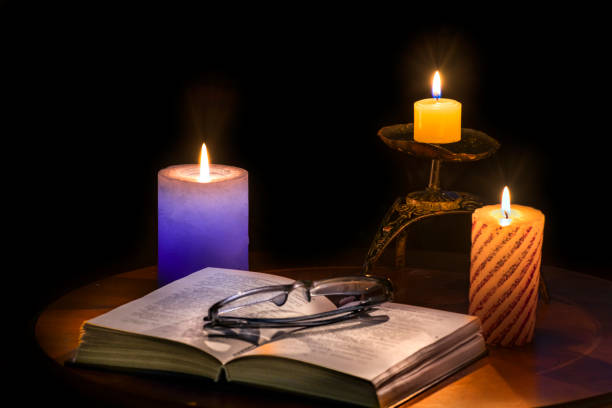 Opened text book with eyeglasses, near are glowing candles, all things on a dark background, concept of old reading fashion