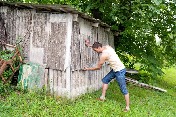 Photo of Young man farmer in garden standing fixing building shed in green summer in Ukraine gray slate wall