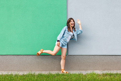 Full length portrait of happy excited beautiful woman in casual jeans denim style in summertime standing near green and light blue wall, looking at camera greeting and waving her hand and surprised.