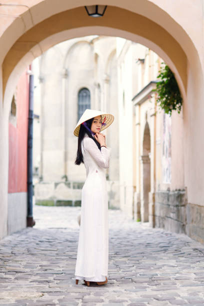 Happy asian girl dressed in traditional Ao Dai white dress with vietnamese conical hat Non La, Leaf Hat. Happy asian girl dressed in traditional Ao Dai white dress with vietnamese conical hat Non La, Leaf Hat. Asian girl walks on the street at cityspace background. ao dai stock pictures, royalty-free photos & images